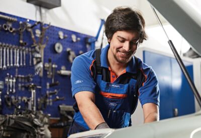 Mann in blauer Latzhose arbeitet in offener Motorhaube in KFZ-Werkstatt