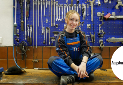 KFZ Mechanikerin Tanja Kennerknecht sitzt im Schneidersitz auf Werkbank vor Werkzeugwand